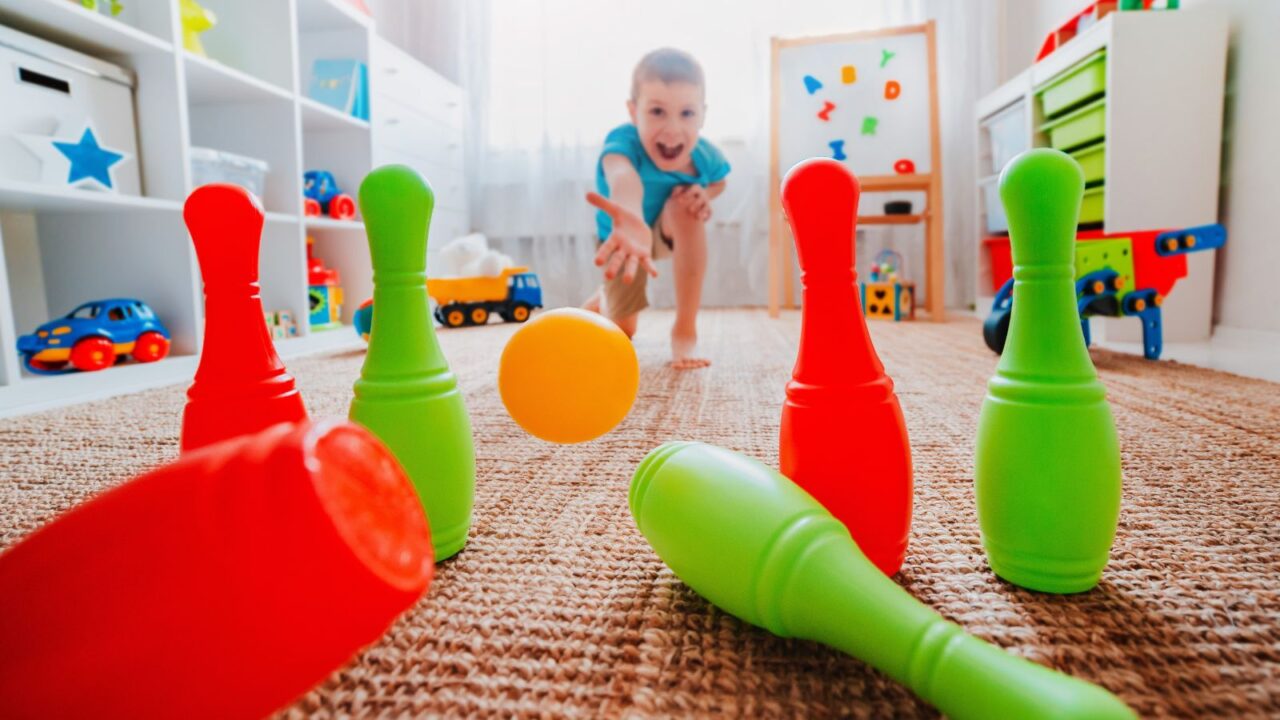 A boy throws ball into a home bowling alley and smashes the bowling pins. A concept of active play in the home room.