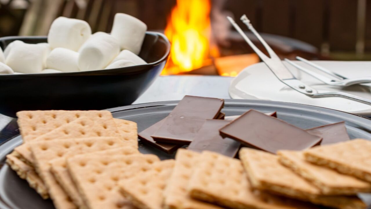 Ingredients to make smores. Graham crackers blurred in foreround. Chocolate in focus. Marshmallows and campfire blurred in background.