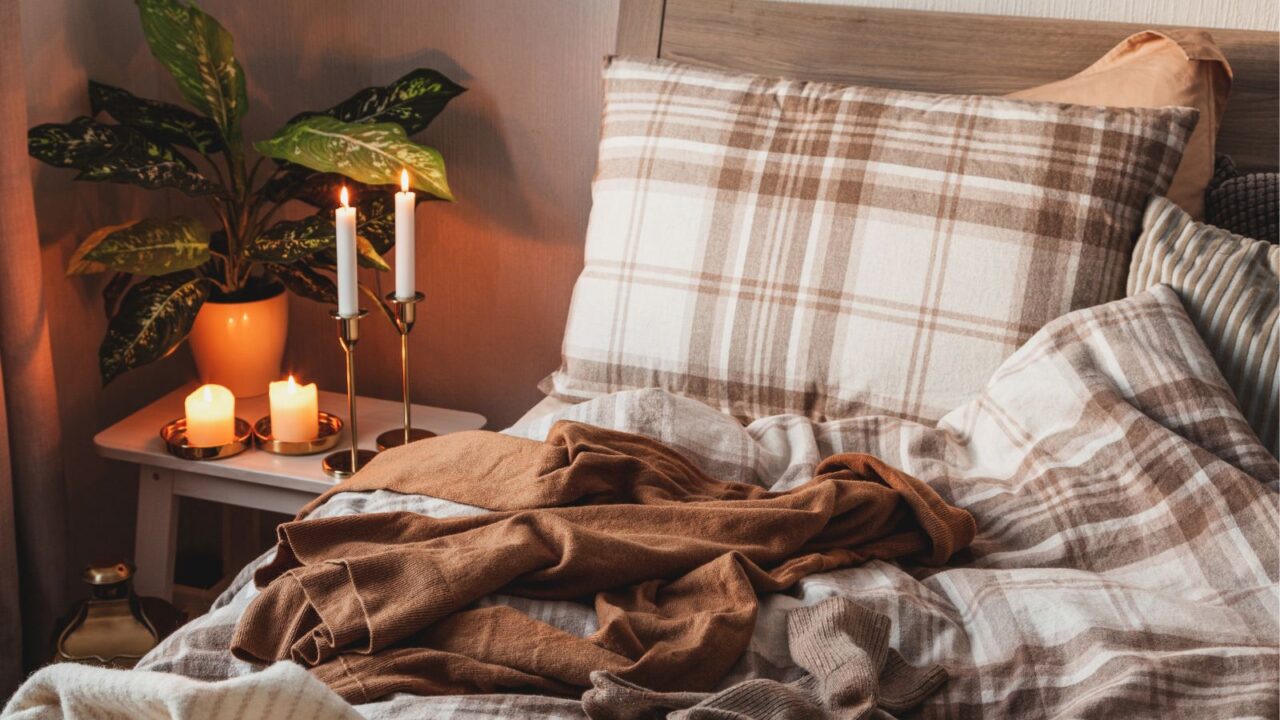 A messy bed with plaid bedding and sweater on it, and burning candles on the side table.