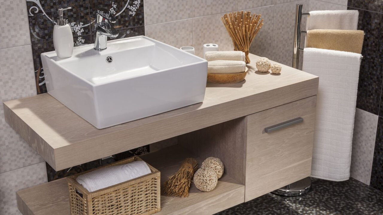 A bathroom interior with a wooden floating vanity with storage basket, a white vessel sink with soap dispenser and chrome faucet, folded towels, and a towel rack beside it.