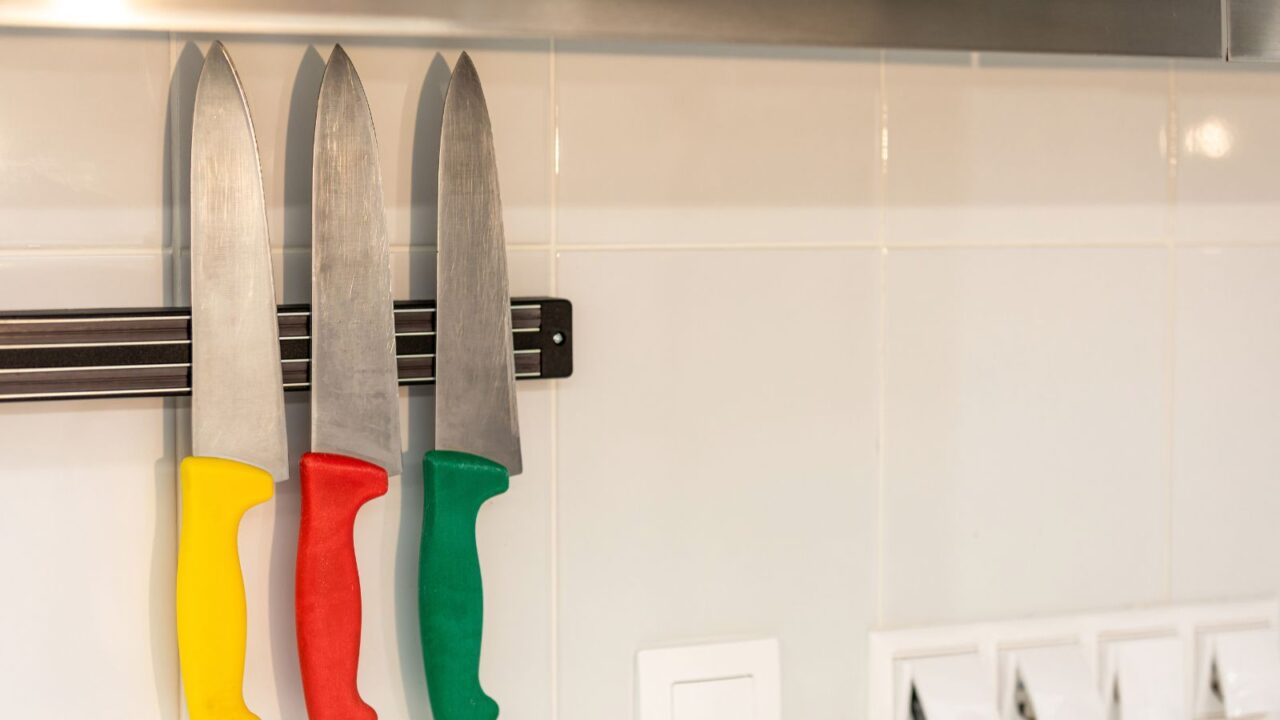 Set of three multi-colored kitchen knives on a magnetic strip on a white wall with sockets nearby.