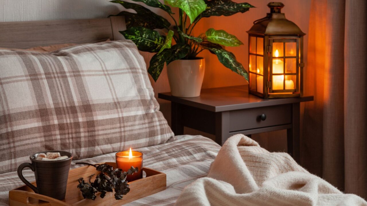 scented candle and a mug on a wooden tray in bed and a lamp and plant are on the side table