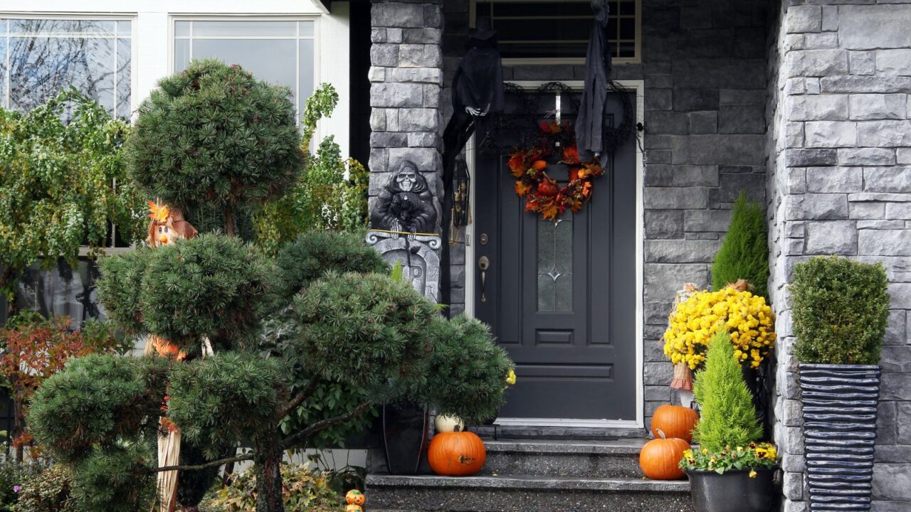 A porch with Halloween decor with gargoyle, hanging witches, wreath, pumpkins, and hidden doll in plants.