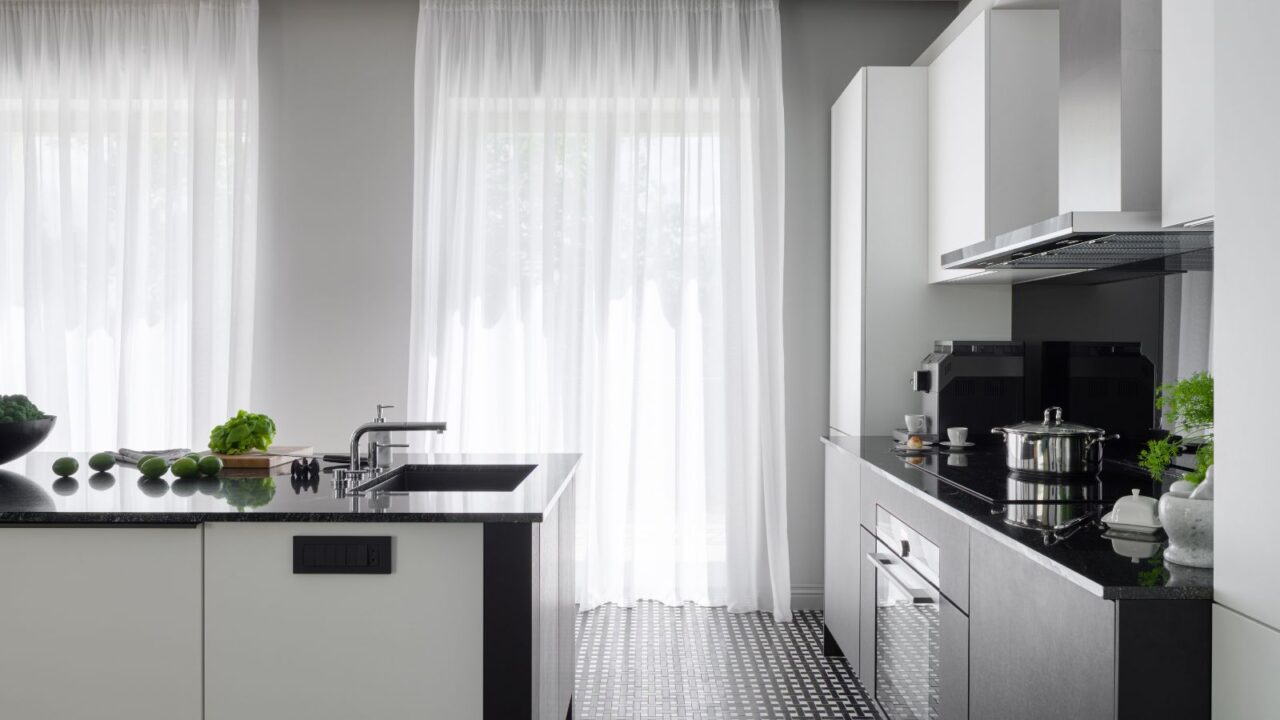 A modern kitchen interior with black kitchen island with sink, black and white cabinets, a black countertop with coffee maker and potted plants, and white sheer curtains.