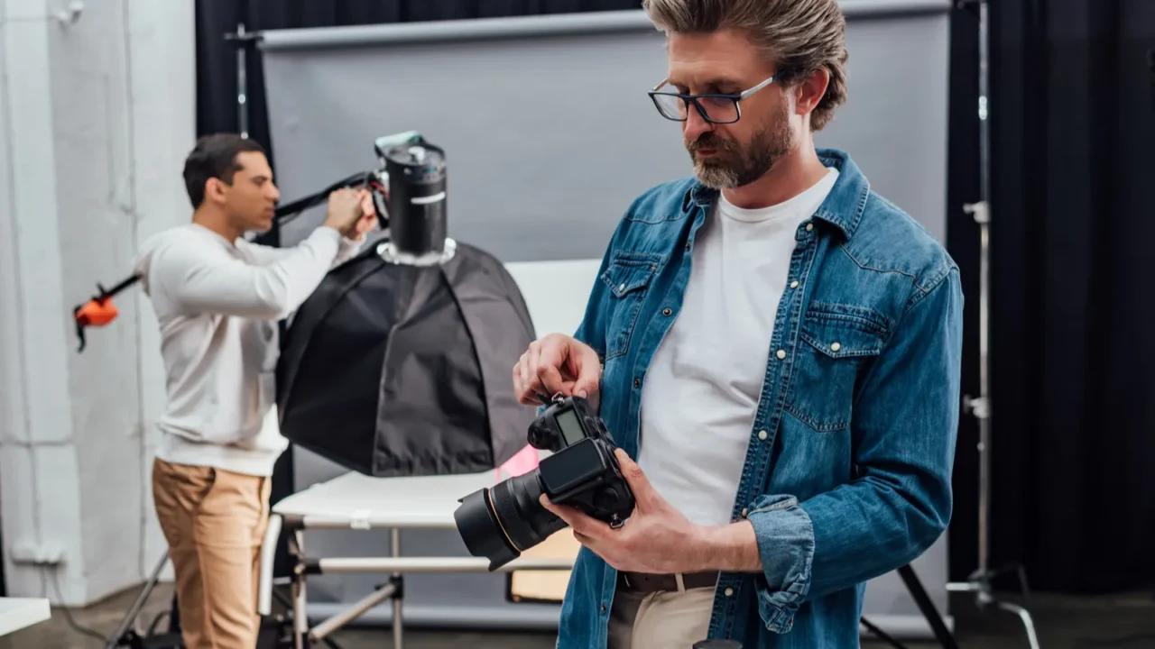 bearded photographer holding digital camera near laptop on table