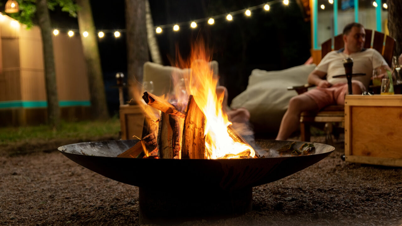 Friends sit near camp bonfire stove against light bulb garland and trees.