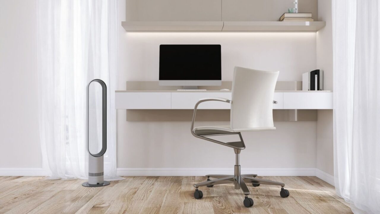 Built in beige working desk with computer, shelf, leather office chair, air multiplier fan, cream wall, parquet floor in sunlight from window with curtain.