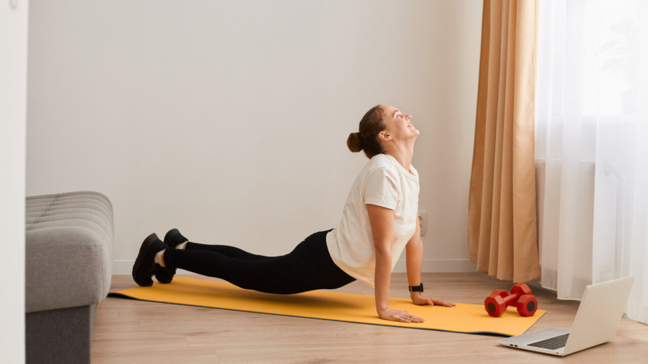 Indoor shot of fit woman making cobra pose on yoga mat, lying on her belly, places her palms under the shoulders, helping herself with hands lift the back up, wearing white T-shirt and black leggins.