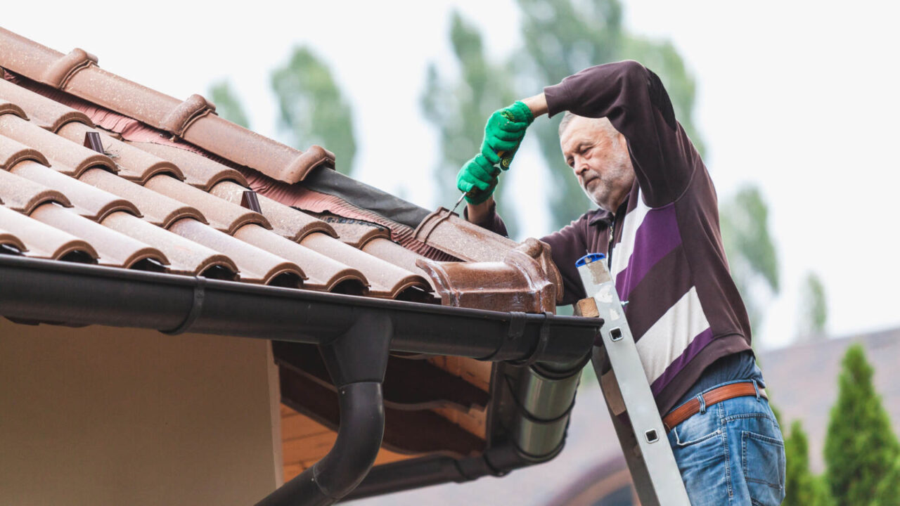 An elderly man repairing the loose shingles of his roof while standing on a ladder