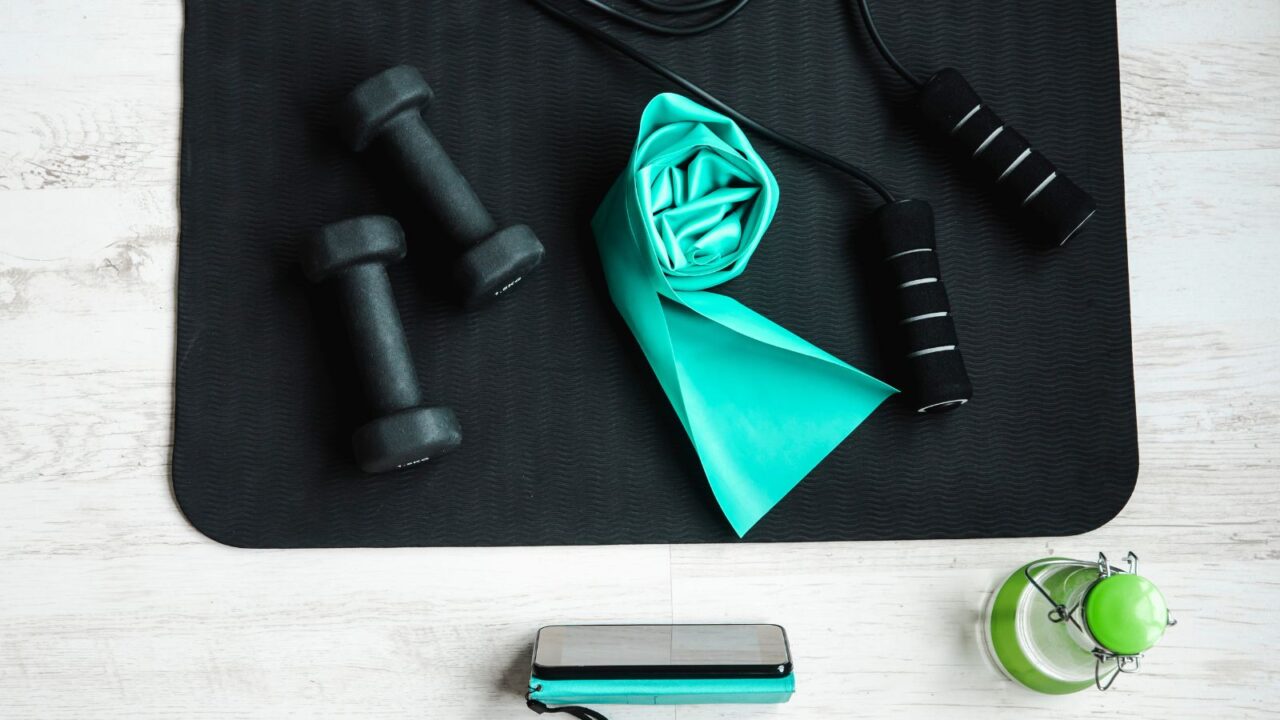 A pair of dumbbells, resistance band, and jumping rope is lying on a black yoga mat