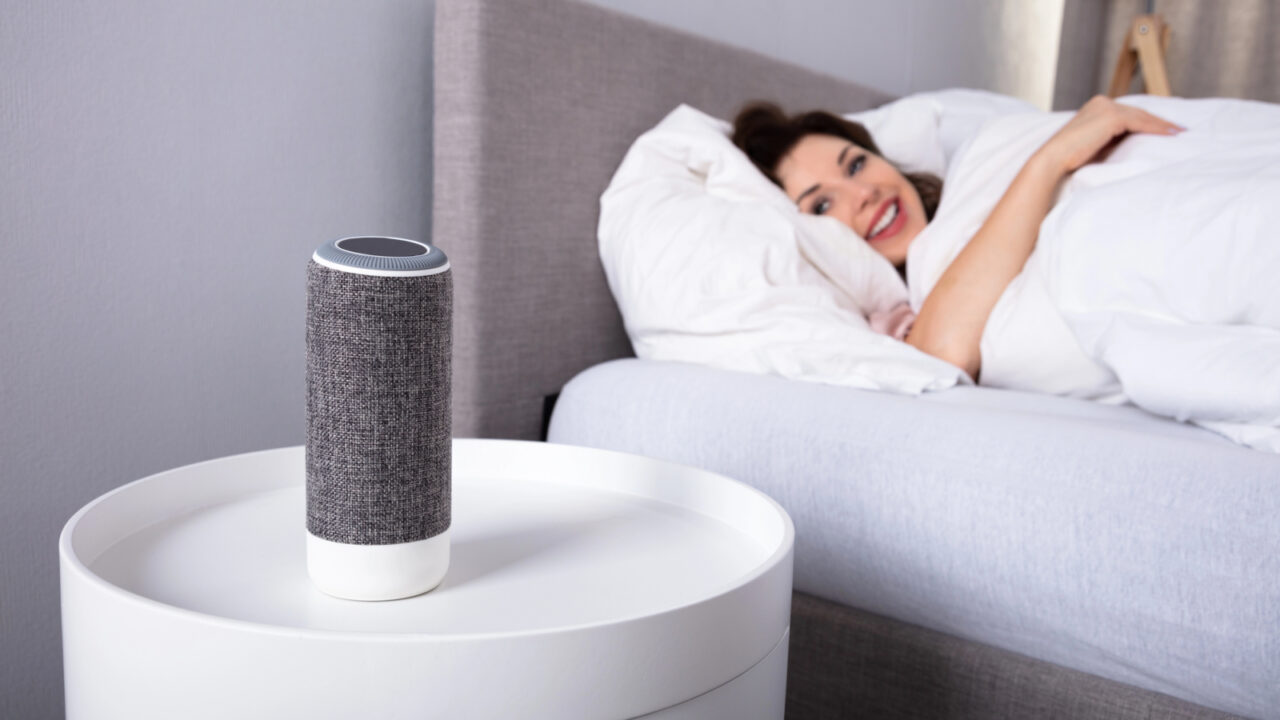 Smiling Young Woman Lying On Bed Listening To Music On Wireless Speaker