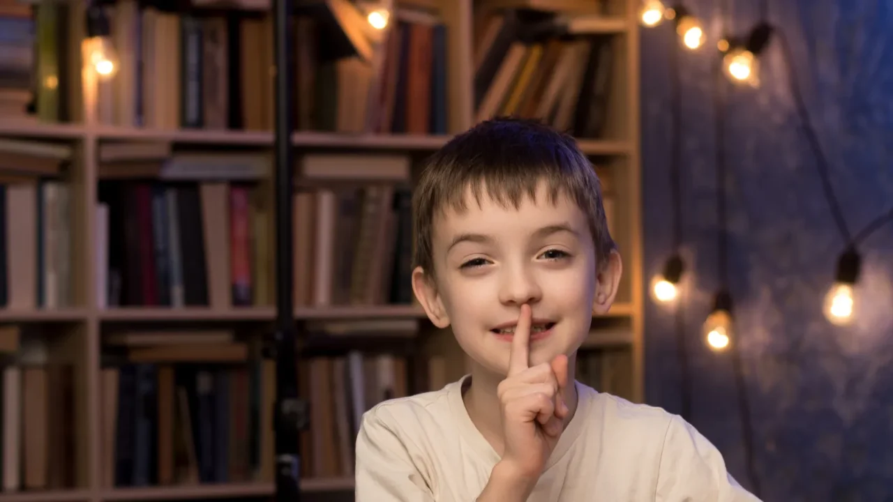 Child in a white shirt sits at a desk, surrounded by books and colored pencils, pondering over a blank paper. Cozy room with string lights and bookshelves in the background. Great for study themes.