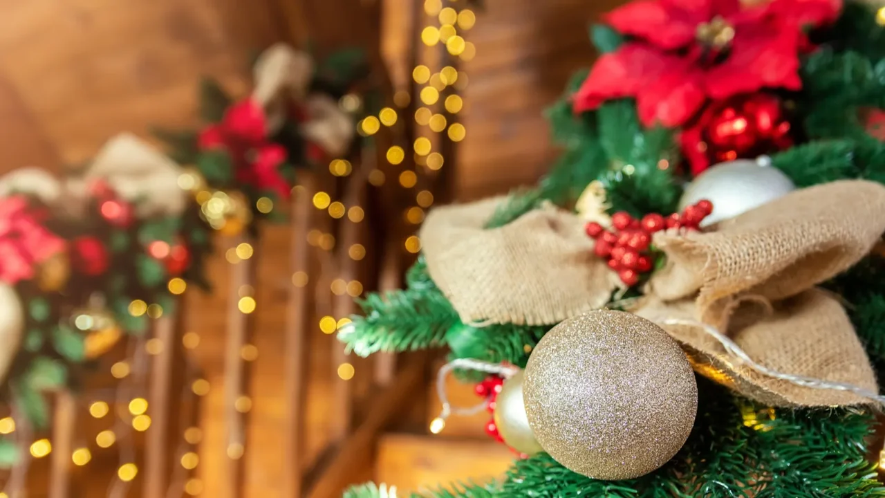 Close-up house wooden staricase handrails railings decorated with artificial holly poinsettia flower, burlap bow, christmas tree golden lights garland. Xmas family home interior decor idea concept