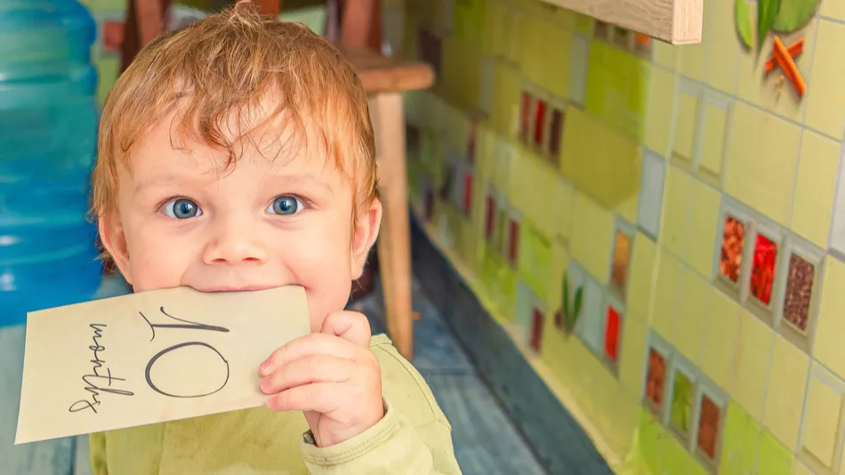 Funny baby in green clothes holds a sign of 10 months old milestone card in his mouth