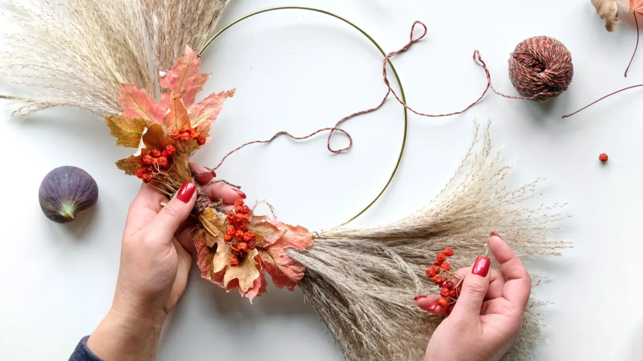 Hands making dried floral wreath from dry pampas grass and Autumn leaves. Hands in sweater with manicured nails tie decorations to metal frame. Flat lay on white table, sunlight with long shadows.