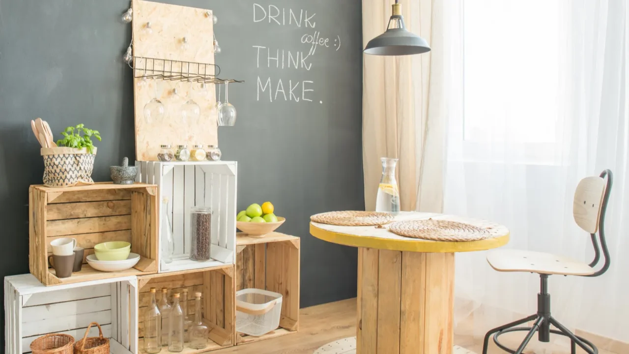 A room with a chair, wooden table, chalkboard wall, lamp, and wooden crates as shelves.