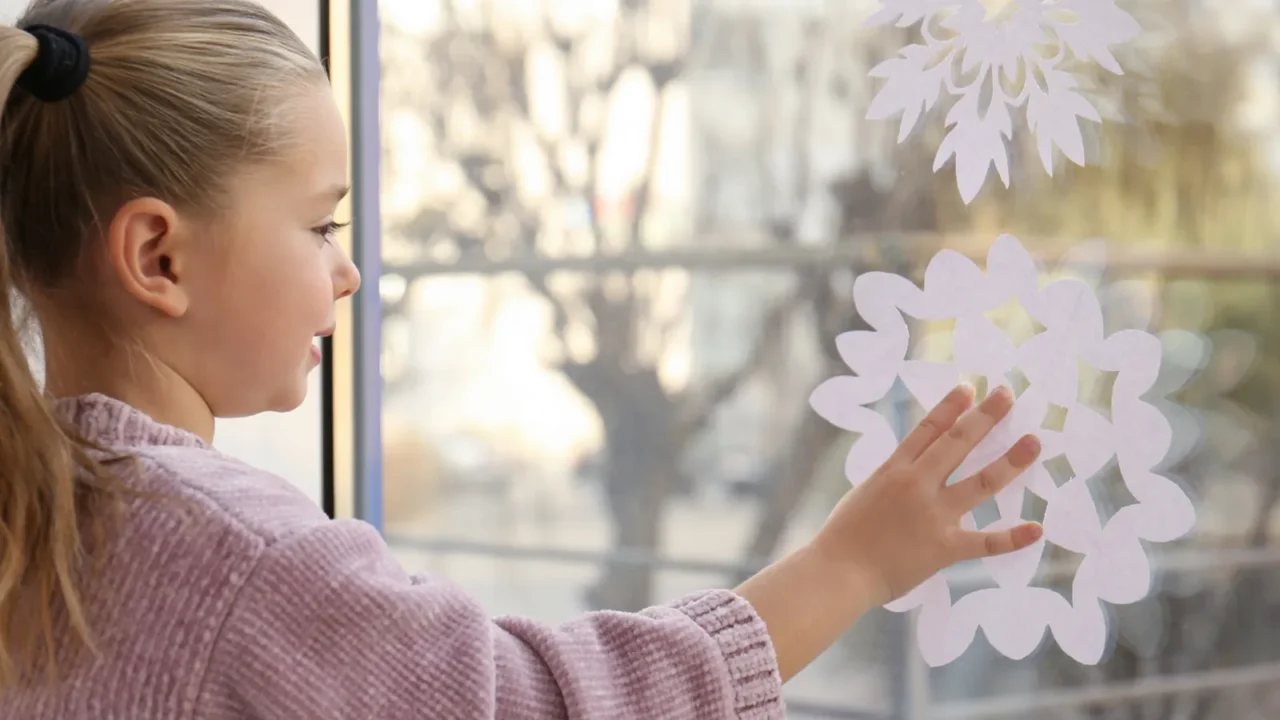 Little girl decorating window with paper snowflake indoors