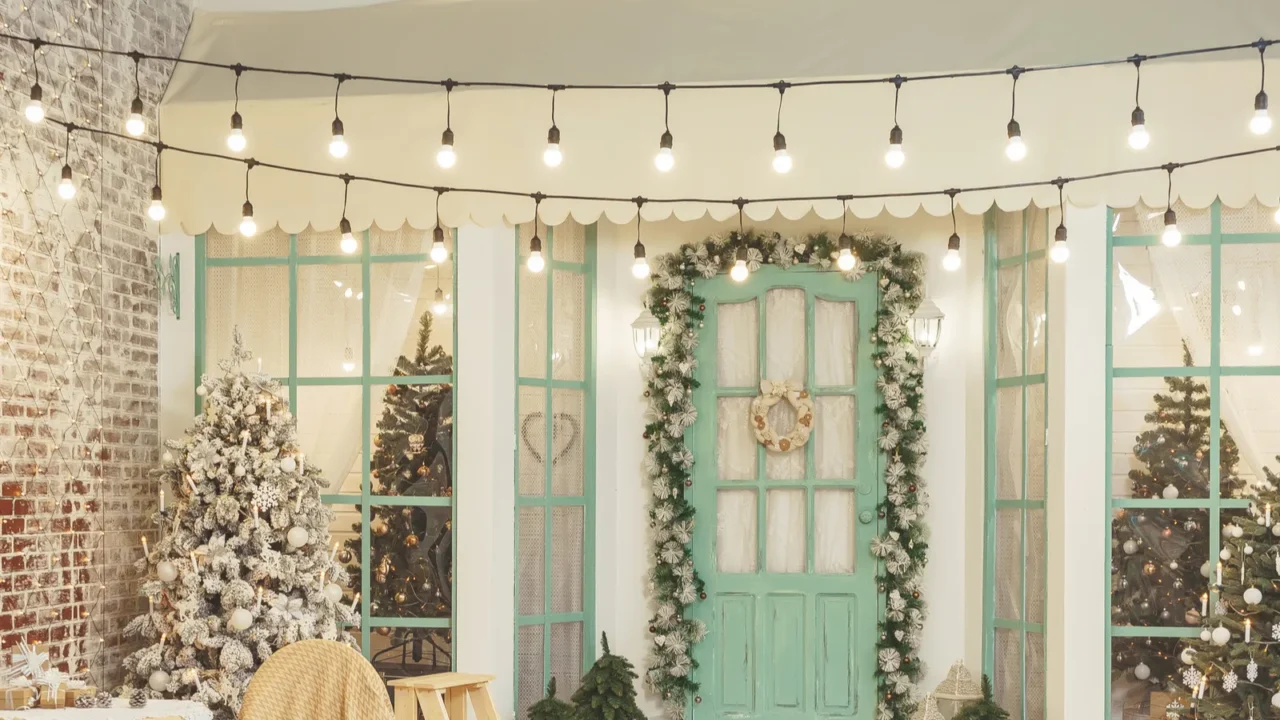 Porch of house with Christmas decorations. Retro street garland with light bulbs over the terrace of house. Garden table and chairs on the winter terrace background of Christmas trees