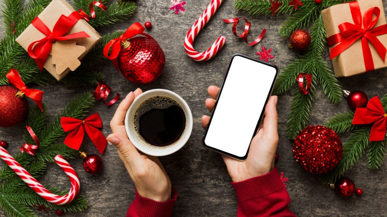 A woman's hands placed on a wooden table, holding a coffee mug and smartphone while surrounded with Christmas decor items, gift box, candy canes, and fir branches