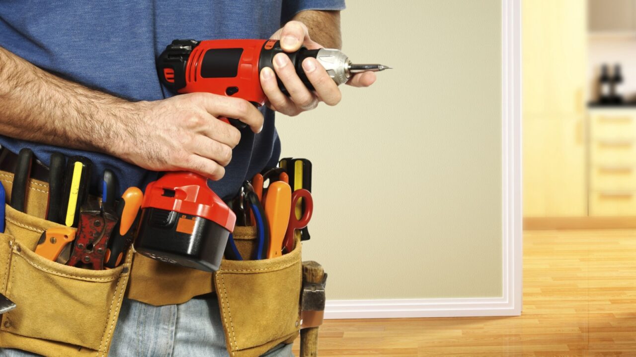 A person holding a red and black cordless drill with a tool belt full of various tools