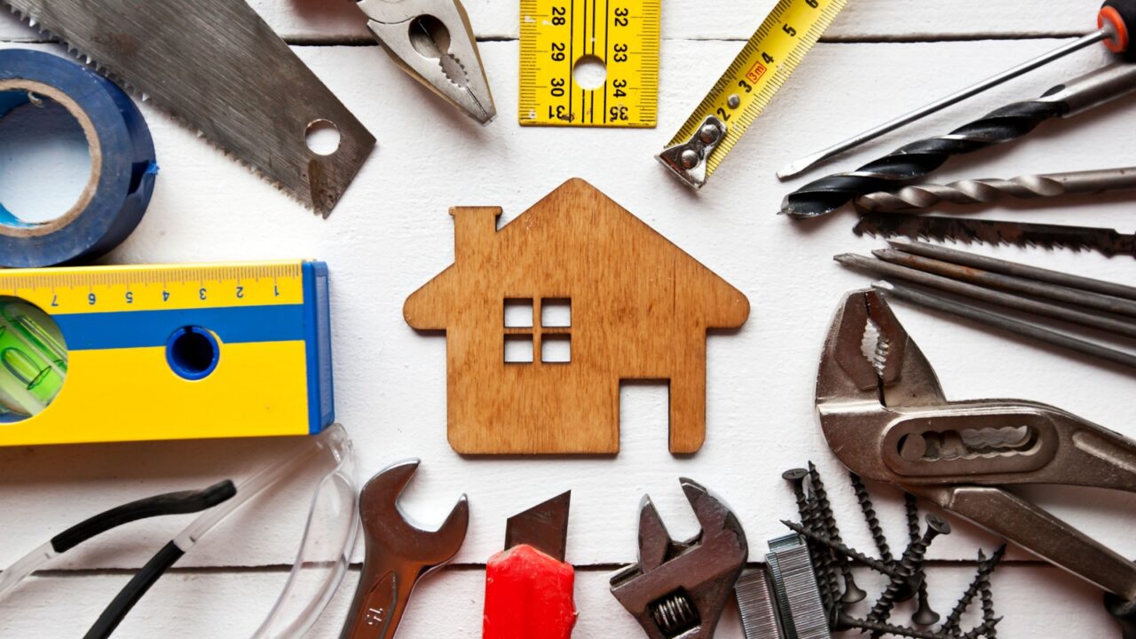 A variety of tools arranged around a wooden house cutout on a white wooden background