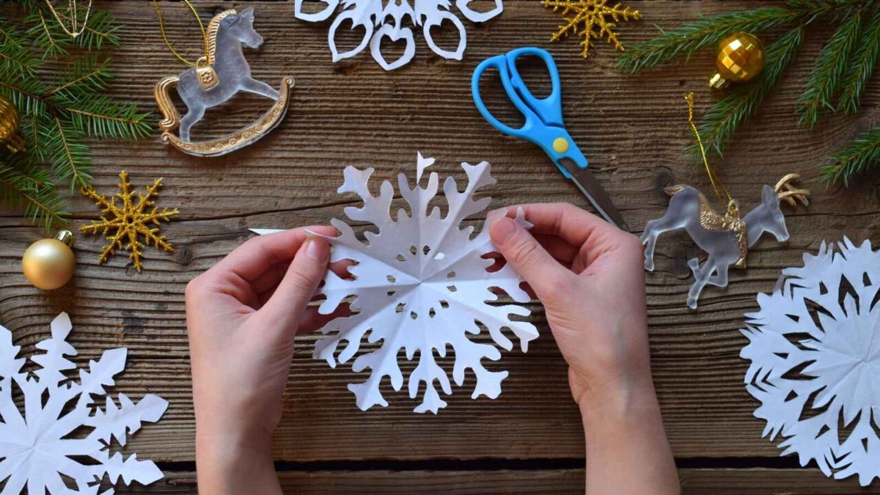 Top view of a man's hand making a paper snowflake on a wooden table with various ornaments, mini figurines and scissors placed across the table