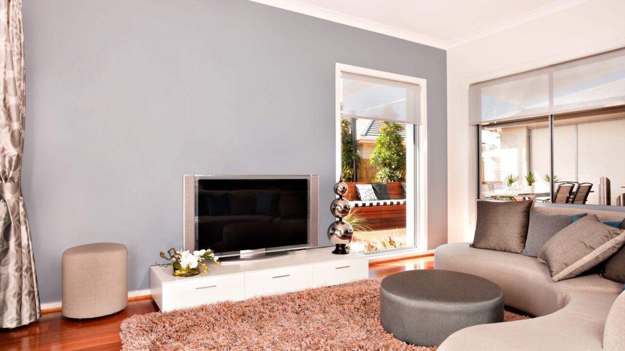 Luxury living room inside decoration using wool carpet on the wooden floor near the grey colour round sofa with pillows and round table near the glass door. There is a led or LCD TV on a grey wall.