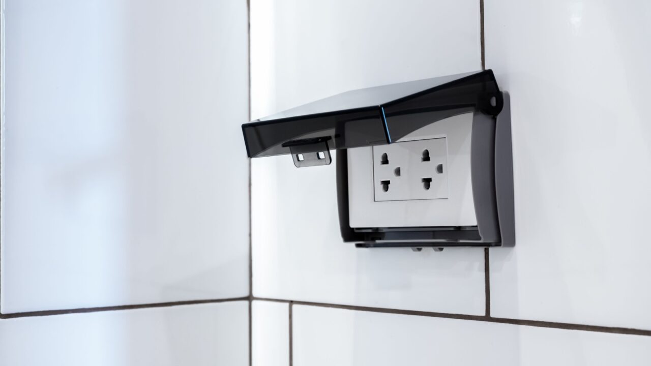 Close-up empty white electrical plug socket plate panel with transparent plastic shield on tiles in bathroom wall.