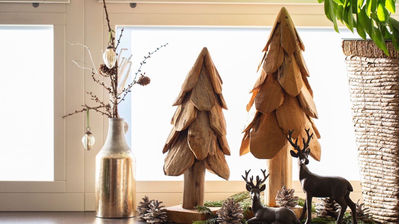 Wooden miniature Christmas trees and reindeers along with pinecones and a vase with dried flowers on a windowsill.