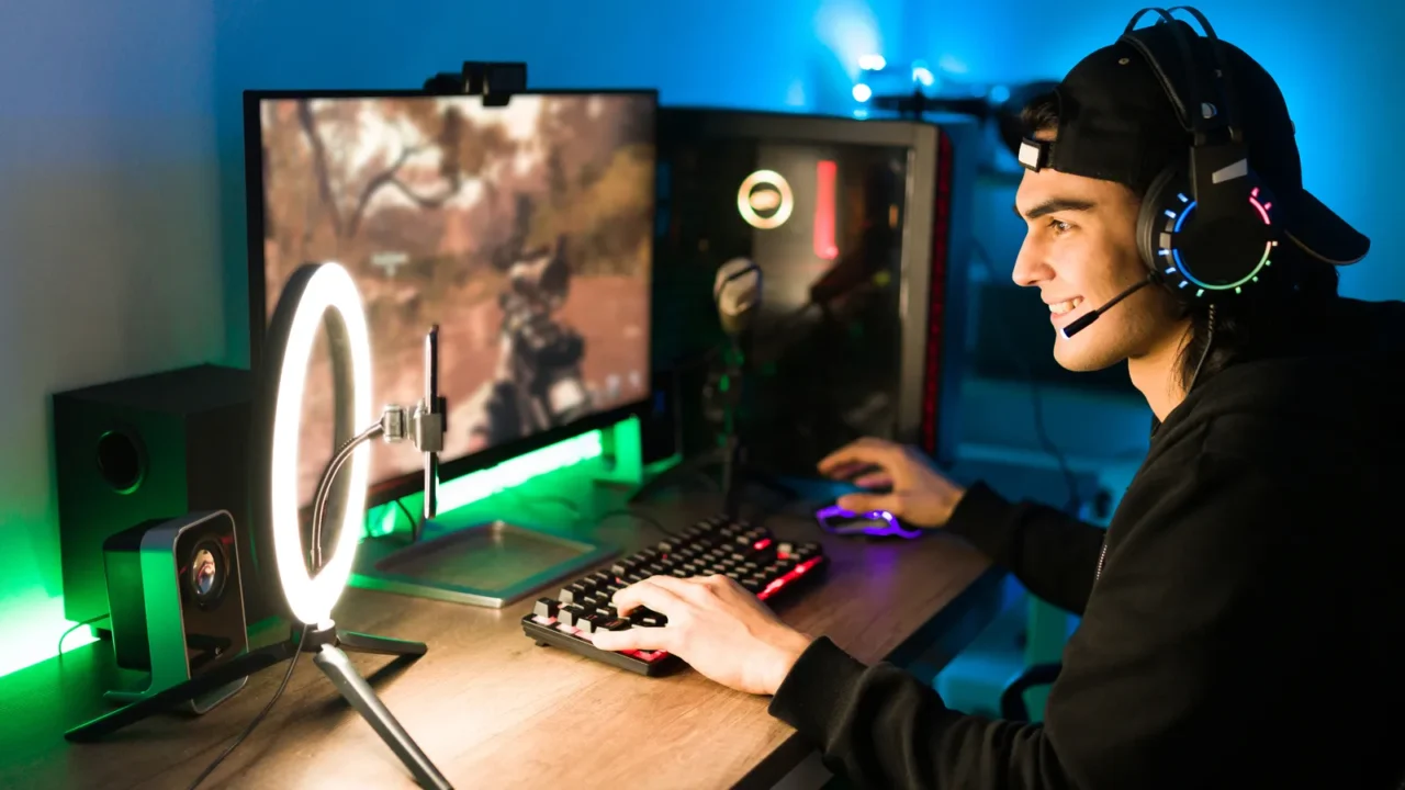 Smiling young man live streaming his online video game using a smartphone and a ring light. Happy gamer ready to start playing in a gaming computer