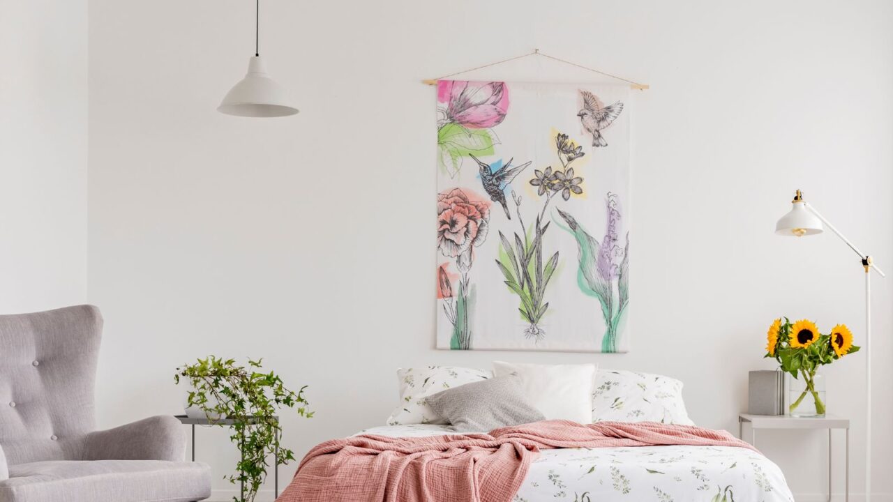 Nature lover's bright bedroom interior with a wall art of flowers and birds painted on a fabric above a bed which is dressed in green plants pattern on white linen. Real photo.