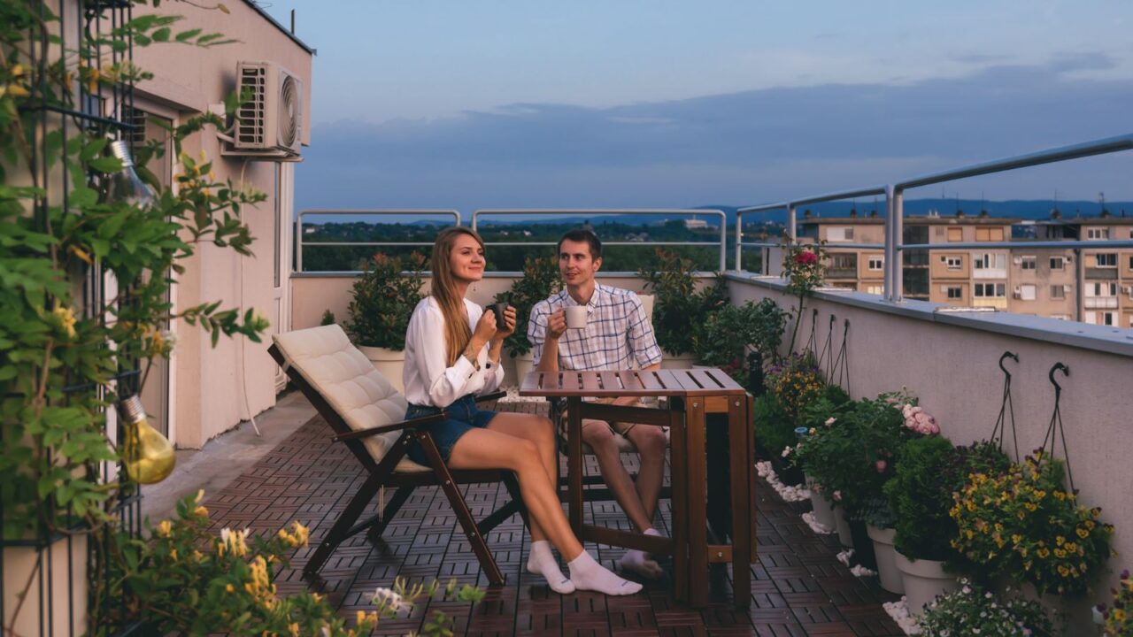 Young couple relaxing outdoors on urban rooftop garden with blooming flowers. Man and woman in casual clothes enjoying sunset and drinking tea or coffee at apartment balcony terrace with city view.