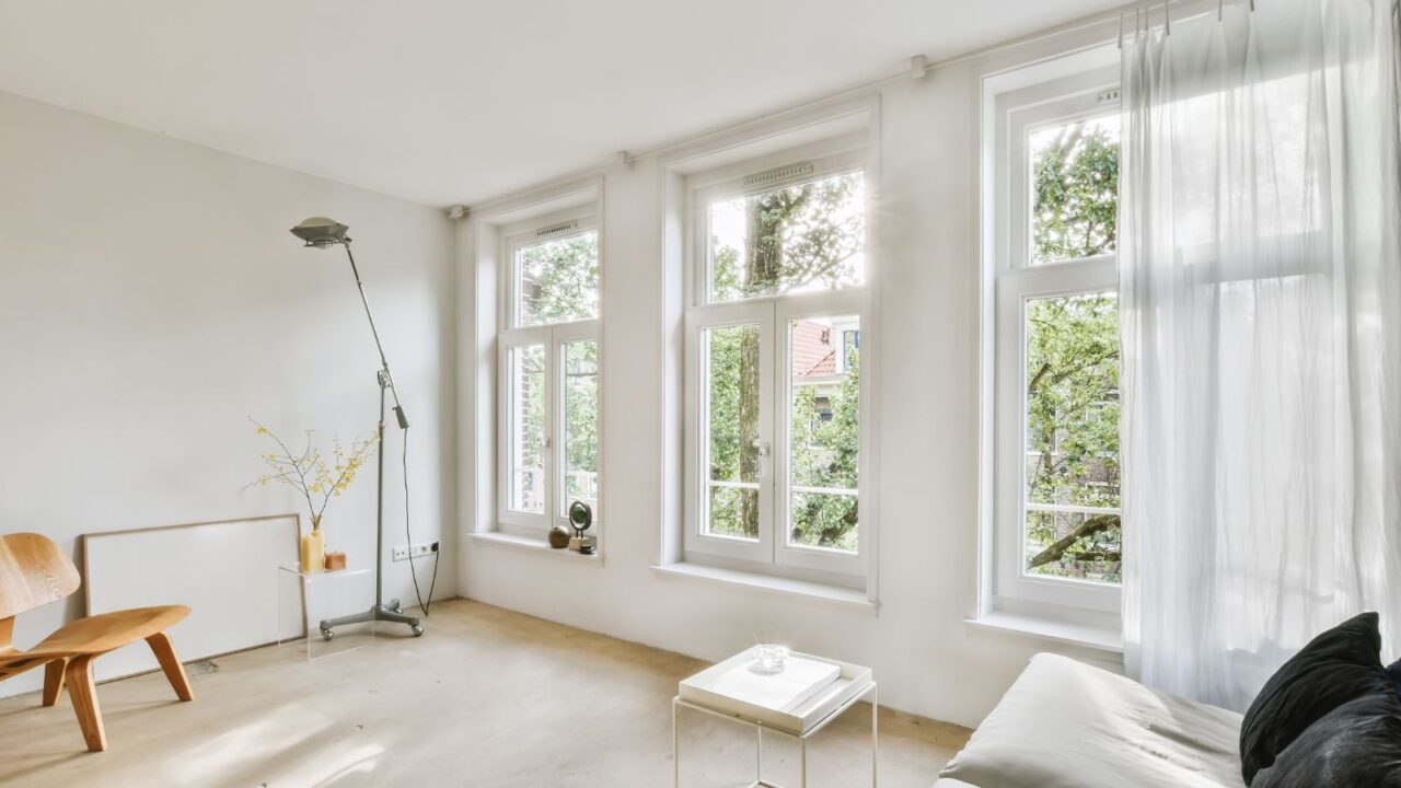 A living room with white walls and wood flooring, two large windows in the wall is covered with sheer curtains.