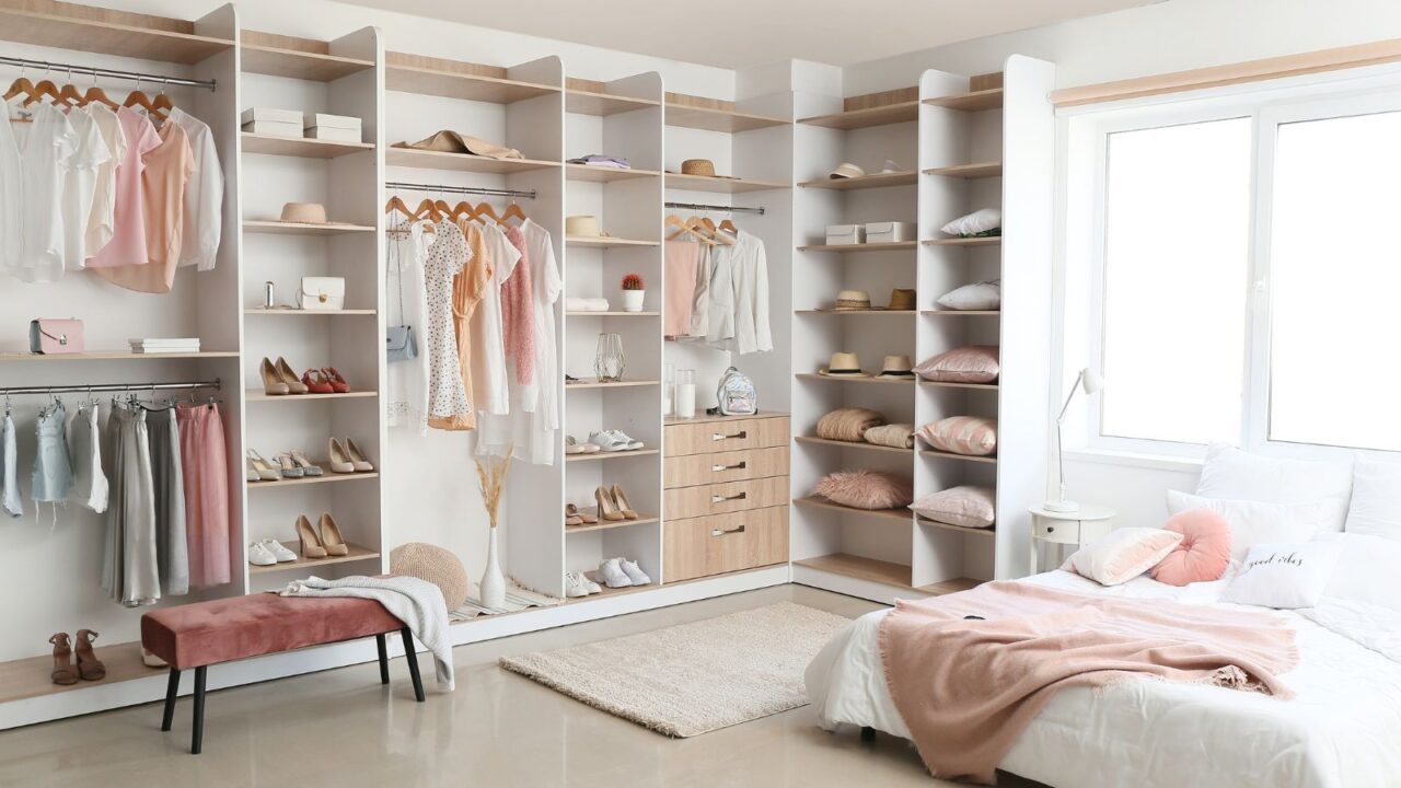 An open plan wardrobe with hanging clothes, shelves with shoes, bags, hats, and storage boxes, a rug, and a comfortable bench.