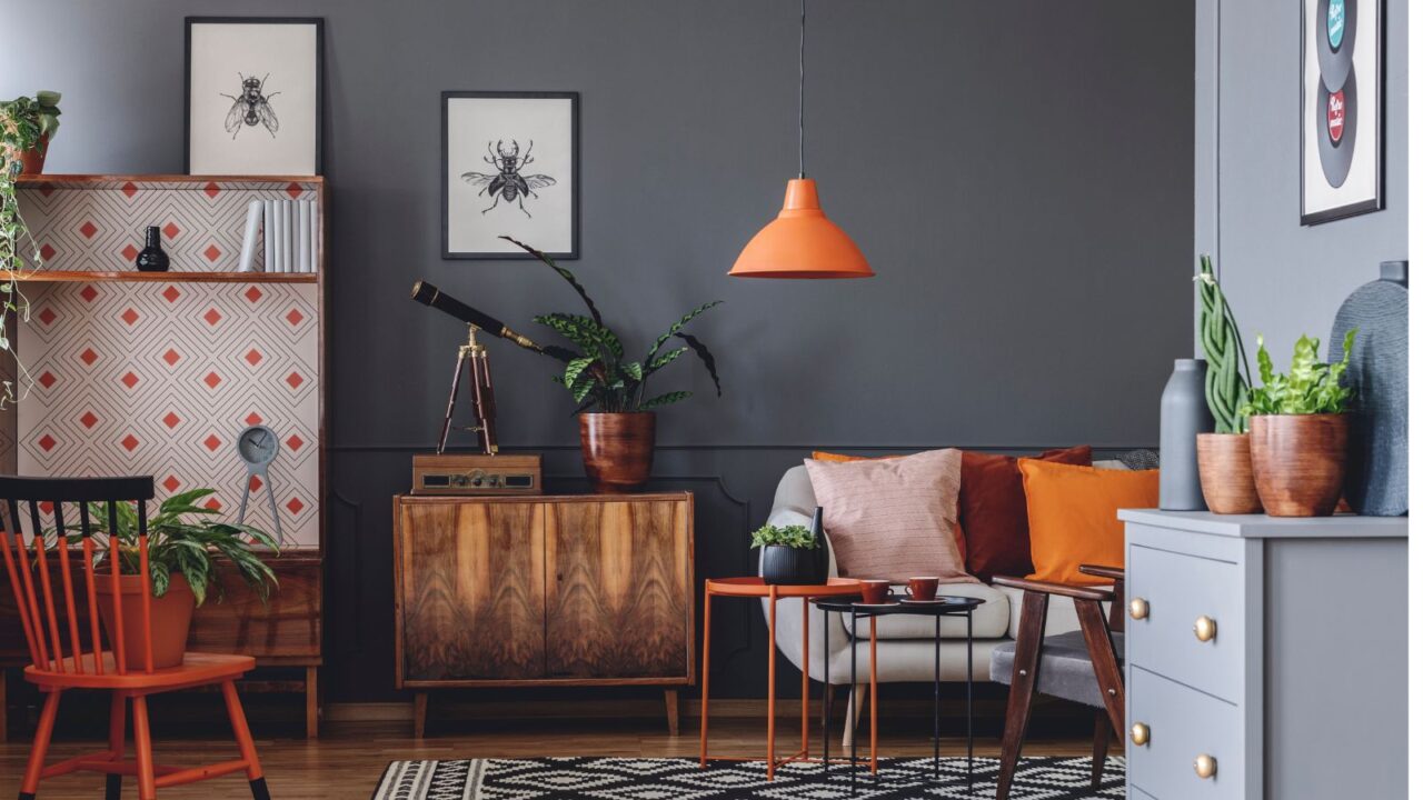 A rustic living room interior with wooden flooring, sofa with colorful throw cushions, nesting tables, a wooden dresser, and houseplants.