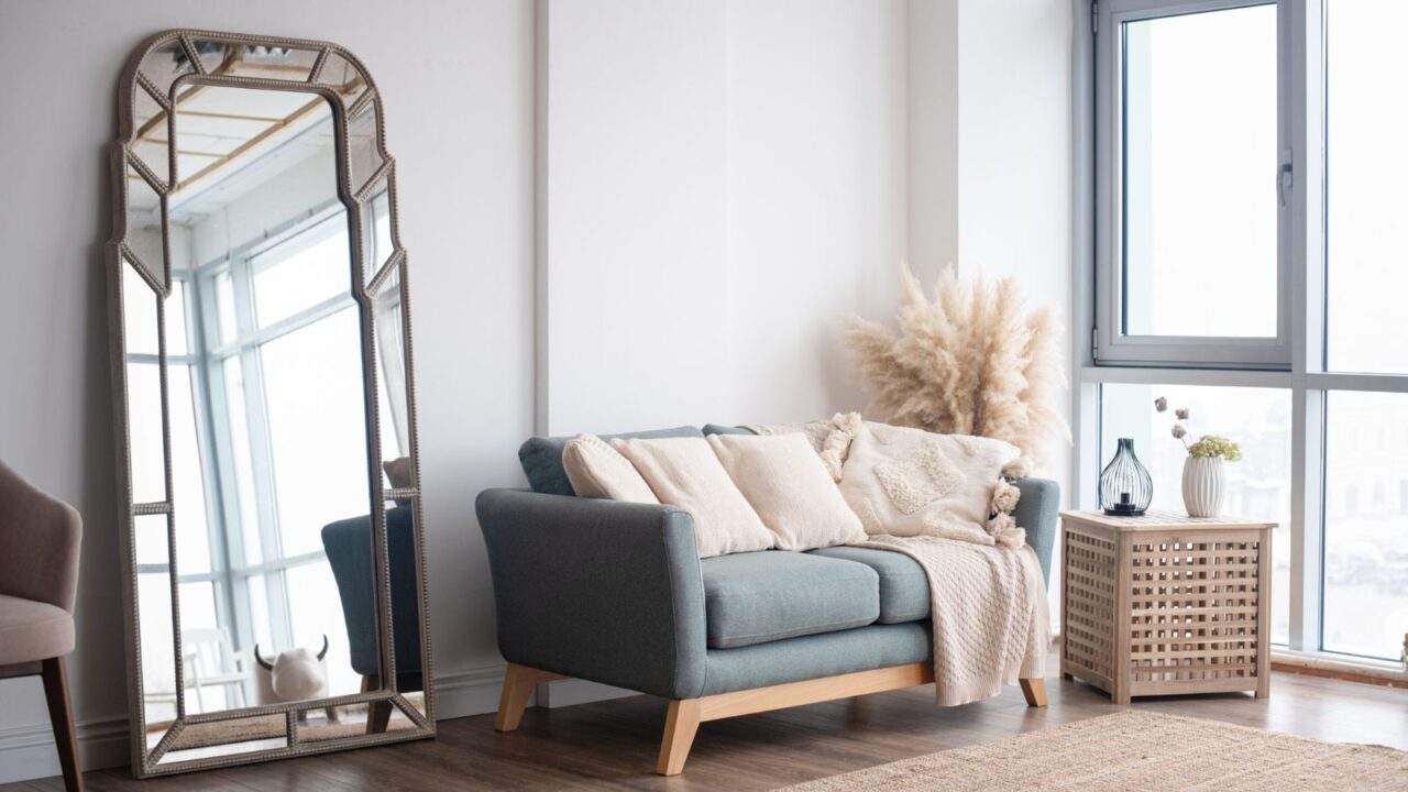 A brightly lit living room interior with a large mirror against the wall, sofa with cushions and throw blanket and a side table with decorative vases.