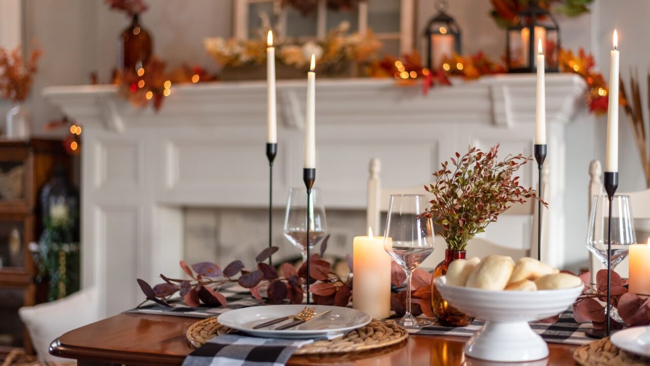 A dining table set for dinner with plates and utensils, candles, dried leaves, and a vase.