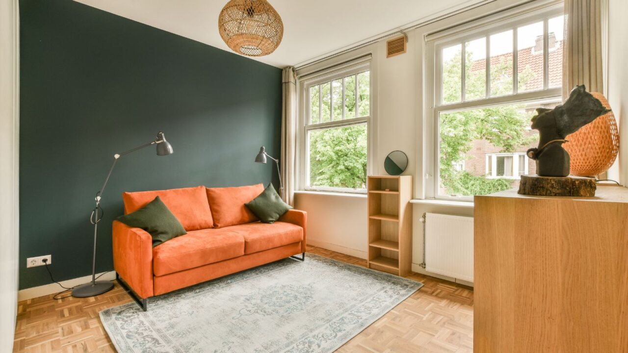 A living room interior with orange sofa in front of a green accent wall, rug, floor lamps, and wooden shelf.