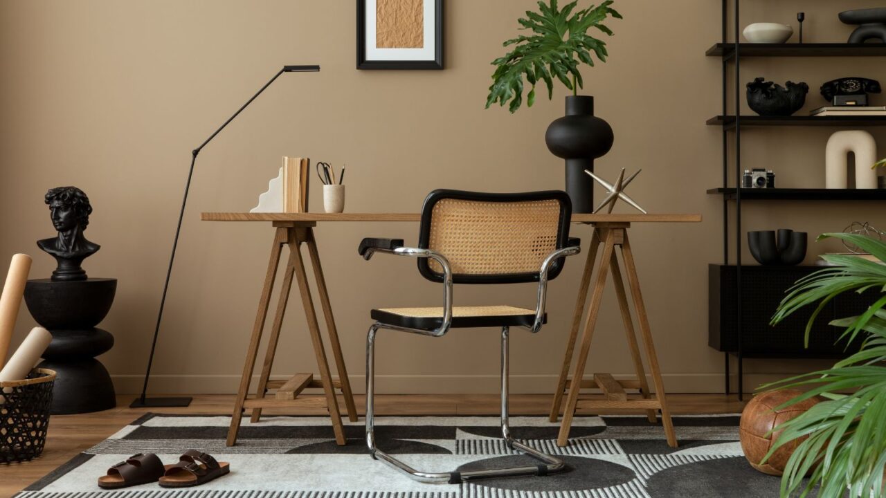 Workplace interior with poster frame, wooden desk, rattan chair, black rack, patterned rug, plant, brown wall, books and personal accessories.