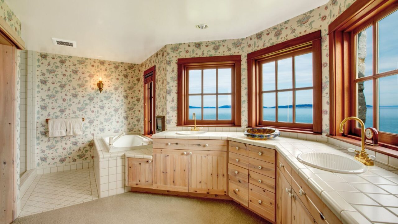 A bathroom interior with vintage floral wallpaper, big windows, bathtub, and carpet.