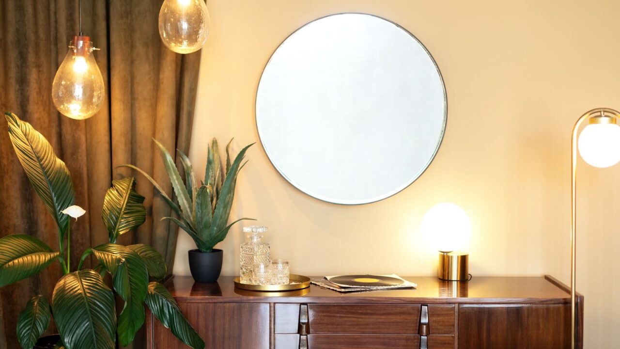 A room with a wooden vanity table with plant pot and a decorative tray, different lamps, and a mirror on the wall.