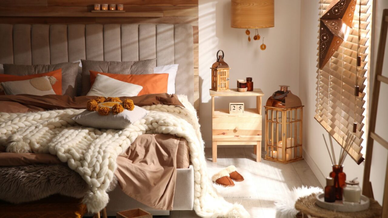 A cozy bedroom interior with a bed with pillows, cushions, and knitted white blanket, wooden side table with decor accents, and a lantern with burning candles.