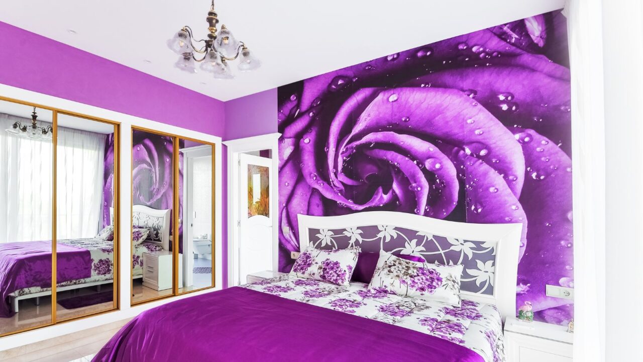 A violet-color bedroom interior with bold wallpaper, wooden flooring, and floral bedding.