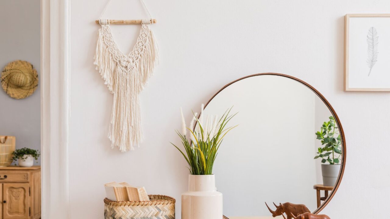 Sunny boho interiors of apartment with macrame, mirror, dressing table, furnitures, flowers, plants, rattan box, books, sculpture, and design accessories. Stylish home decor.