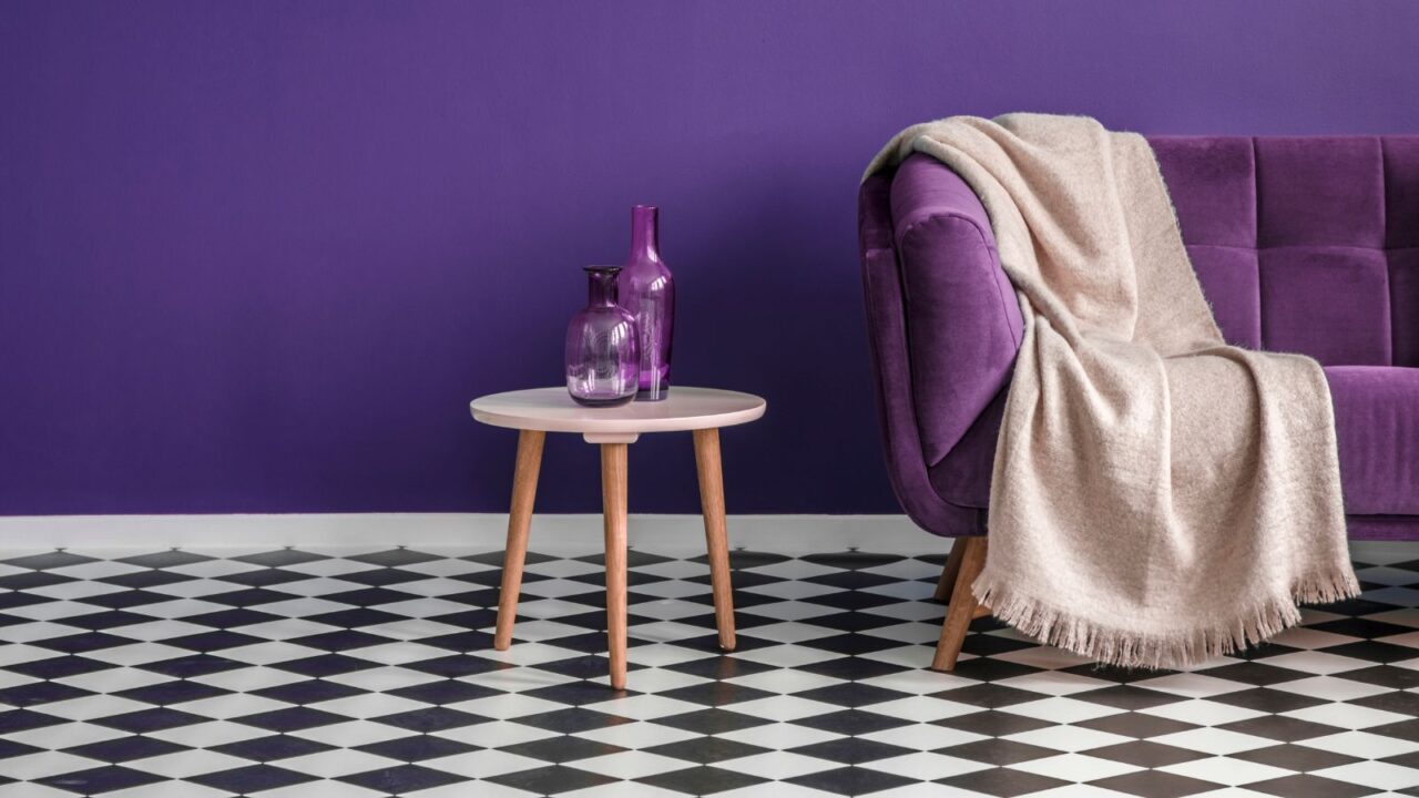 Dark purple sofa with a blanket beside a small table with bottles standing on black and white checkerboard floor in a minimalistic living room interior.