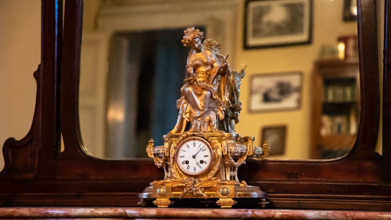 French vintage clock on a table with mirror at the back. Reflection of a bookshelf and frames hanging on the wall can be seen in the mirror.