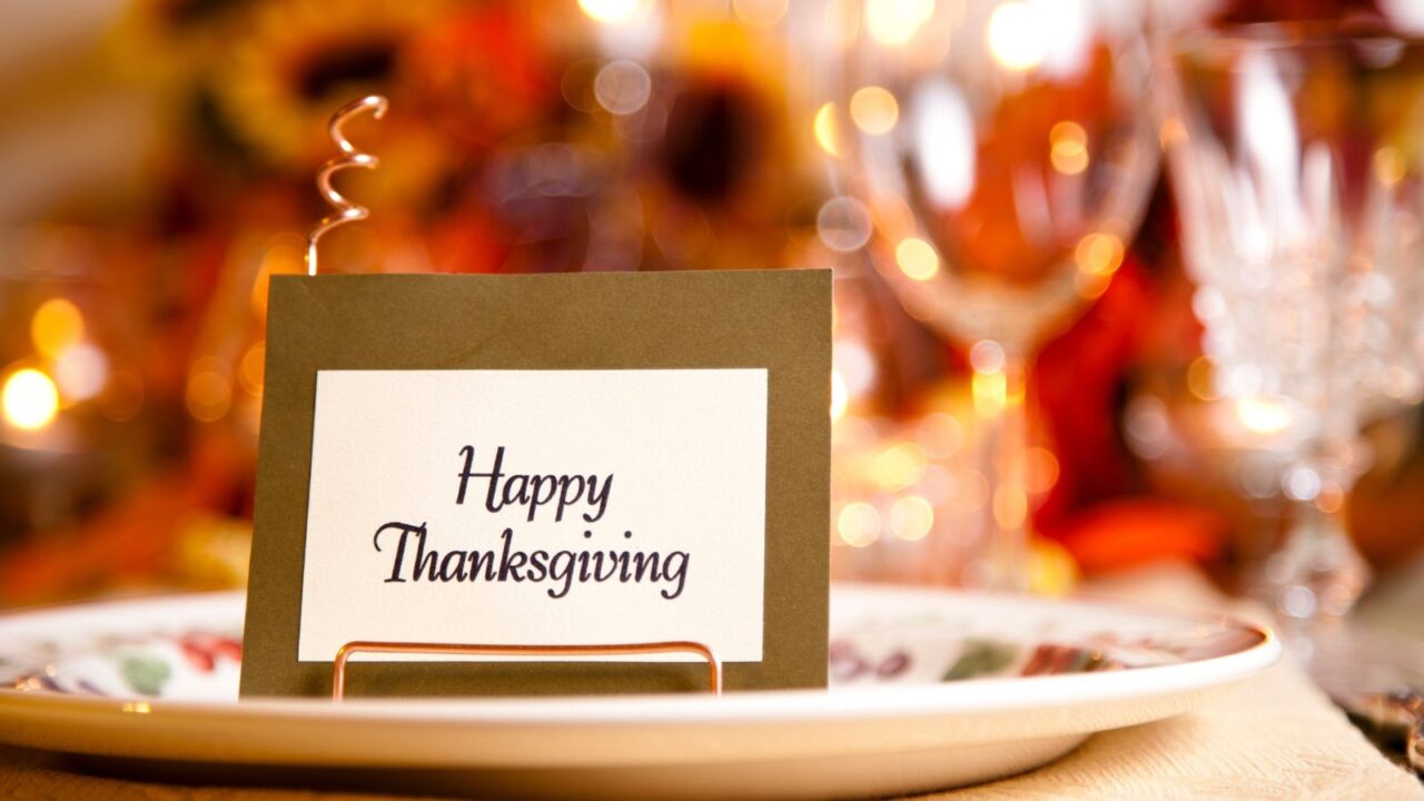 Thanksgiving place card, Happy Thanksgiving script, copper holder, white plate, beige tablecloth, glassware, candlelight bokeh, warm tones, cozy autumn setting, festive atmosphere.