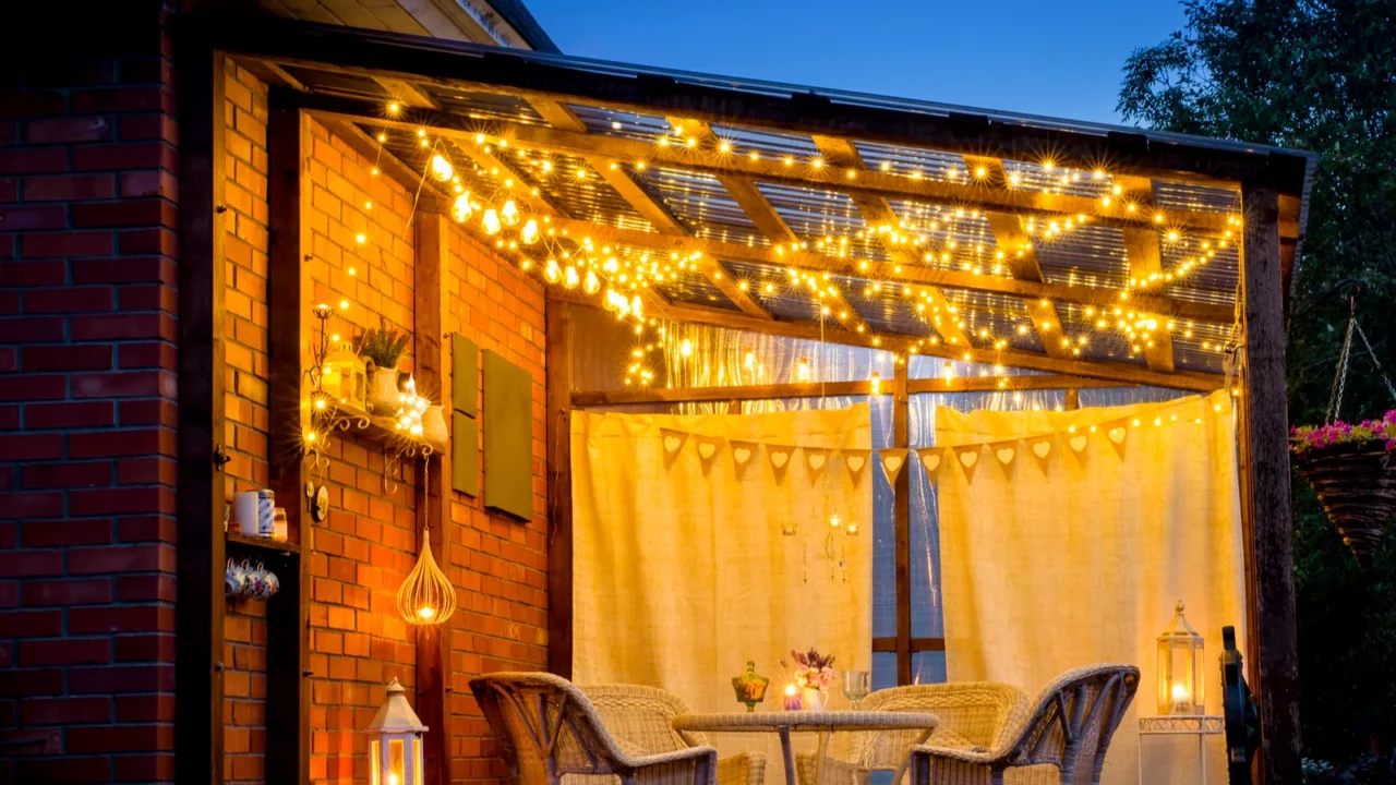 View over cozy outdoor terrace with table and chairs, very romantic lighting, white lanterns, candles burning, led string party lights and bulbs with star effect, in the evening.