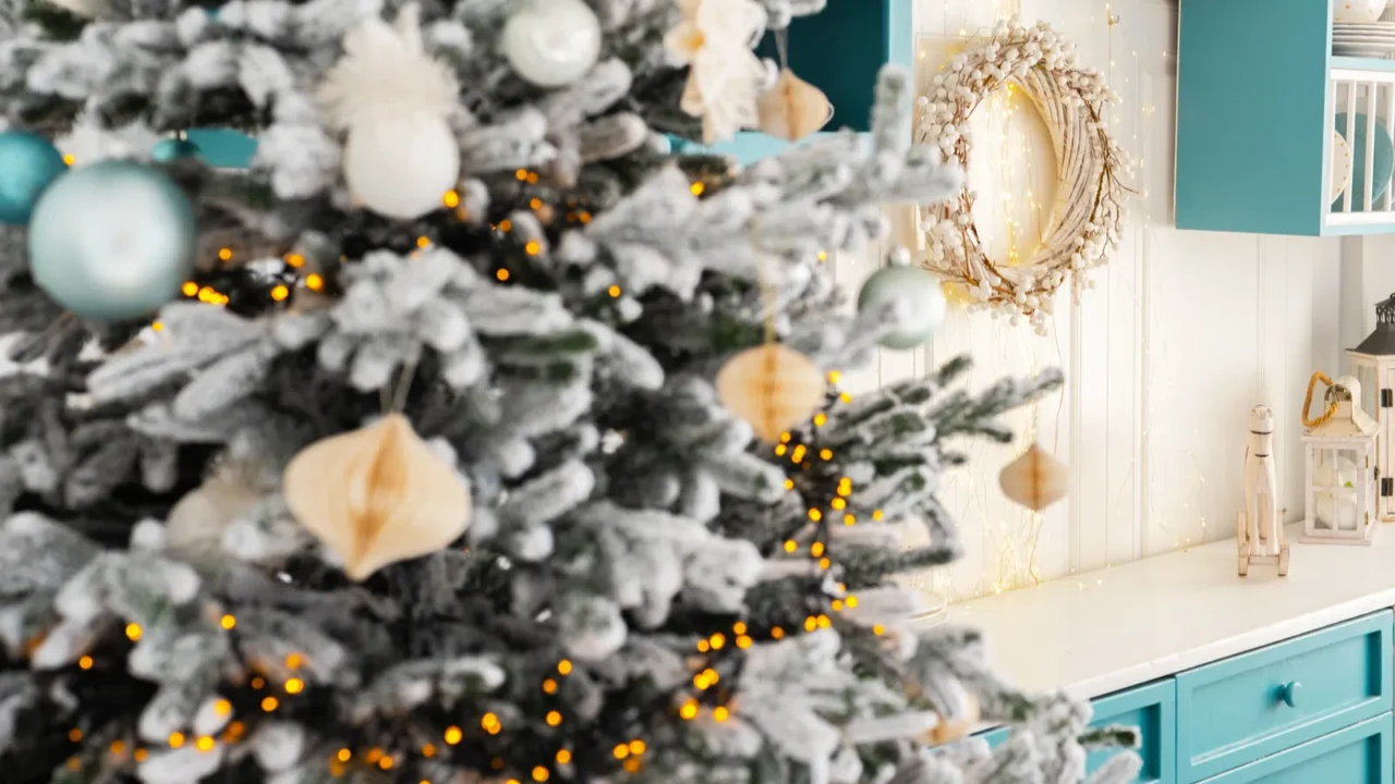 A beautifully arranged living room features a snowy Christmas tree and mint decor. White and mint kitchen at the back with a lantern on the counter and a wreath hanging.