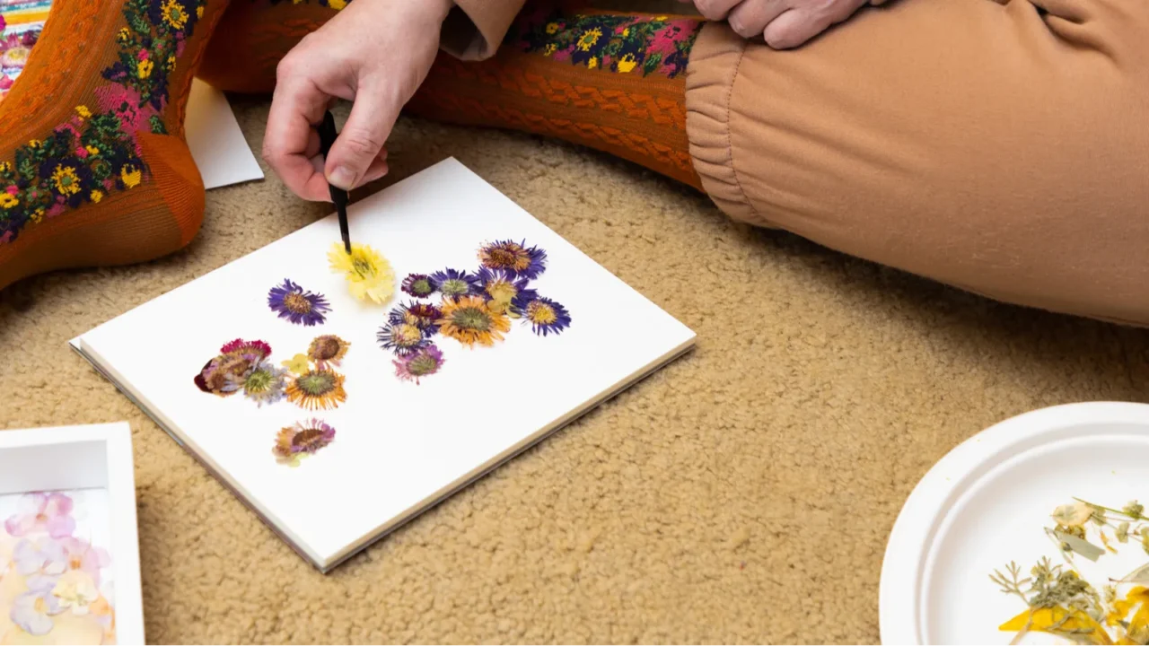 A woman sits on a carpet and makes a pressed flowers frame.